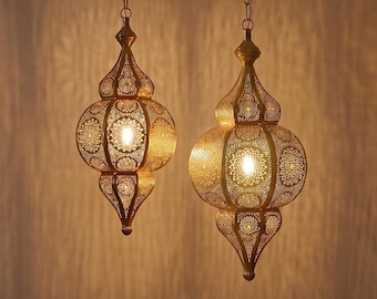 May include: Two gold-toned metal pendant lights with intricate laser-cut patterns hanging from chains. The lamps feature a bulbous, teardrop-shaped design that emits a warm, glowing light, casting detailed shadows on the wall behind them.