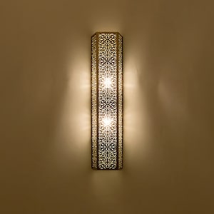 May include: A gold metal wall sconce with a geometric design and three light bulbs. The sconce is illuminated and casting a warm glow.