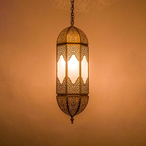 May include: A gold metal hanging lantern with intricate cutouts and a white frosted glass panel. The lantern is lit and casting a warm glow.