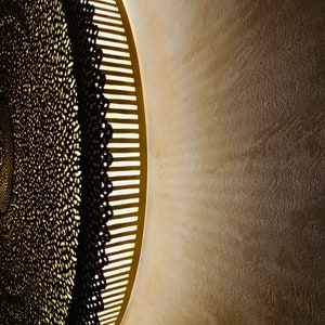 May include: A circular wall light fixture featuring a dark, intricate, lace-like metal overlay with a repeating pattern. The fixture emits a warm, soft glow from behind the metal structure, casting light onto the surrounding wall surface.
