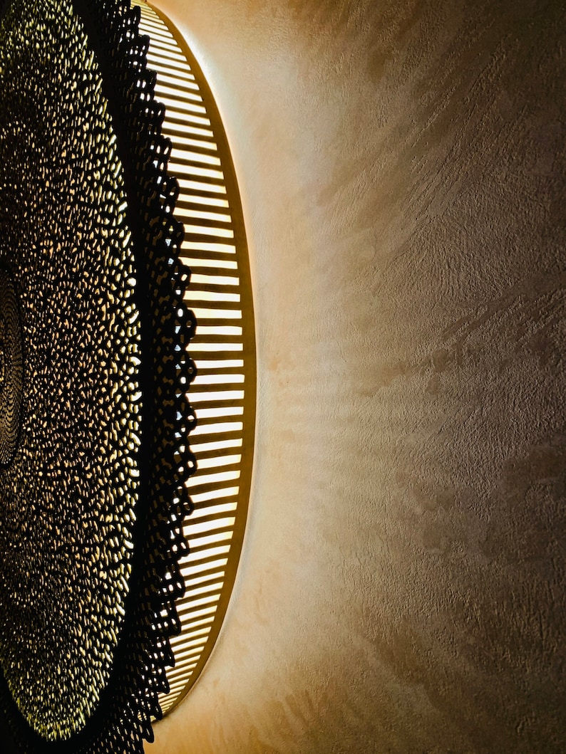 May include: A circular wall light fixture featuring a dark, intricate, lace-like metal overlay with a repeating pattern. The fixture emits a warm, soft glow from behind the metal structure, casting light onto the surrounding wall surface.