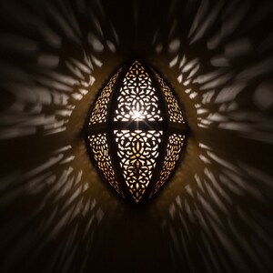 May include: An oval-shaped wall lamp featuring a dark metal frame with intricate, laser-cut filigree patterns. The lamp emits a warm, golden light from within, casting dramatic, elongated shadows in a radial pattern across the surrounding wall surface.