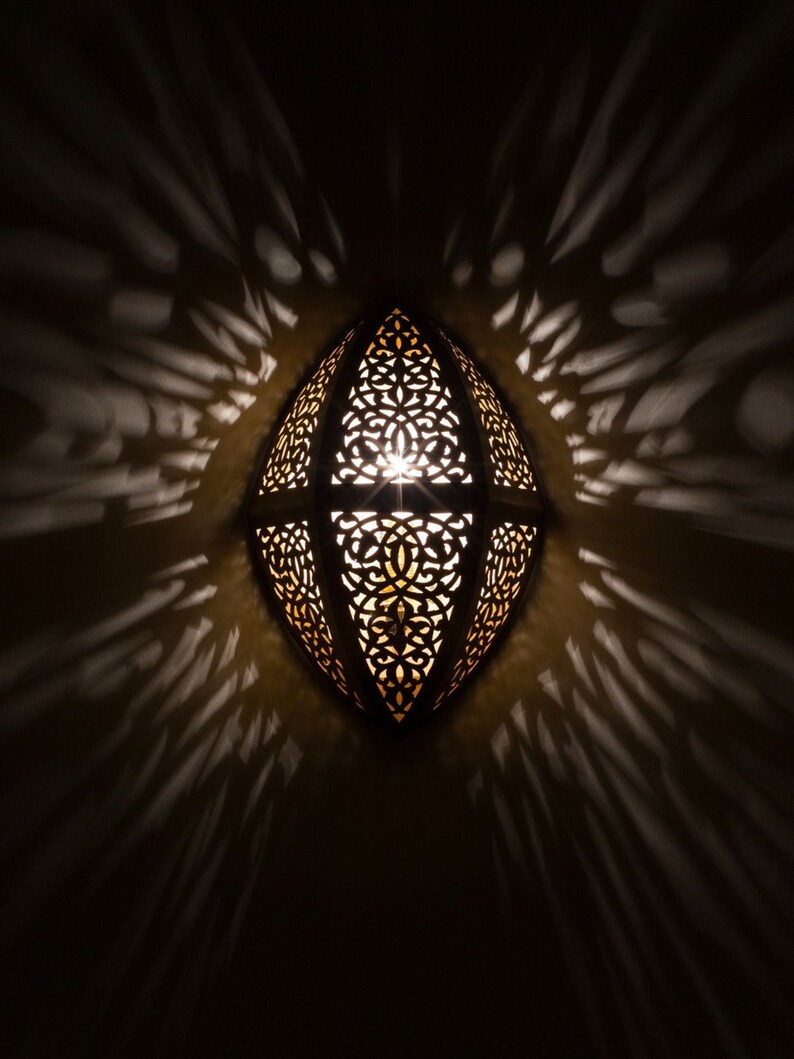 May include: An oval-shaped wall lamp featuring a dark metal frame with intricate, laser-cut filigree patterns. The lamp emits a warm, golden light from within, casting dramatic, elongated shadows in a radial pattern across the surrounding wall surface.
