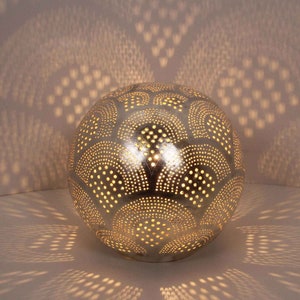 May include: A silver metal lampshade with a decorative pattern of circles and arches. The lampshade is illuminated from within, casting a warm glow.