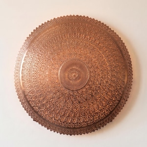 May include: A circular copper-colored wall plaque featuring an intricate, embossed pattern of repeating geometric and floral motifs. The design radiates from a central point toward a scalloped, serrated edge, creating a textured, metallic surface.