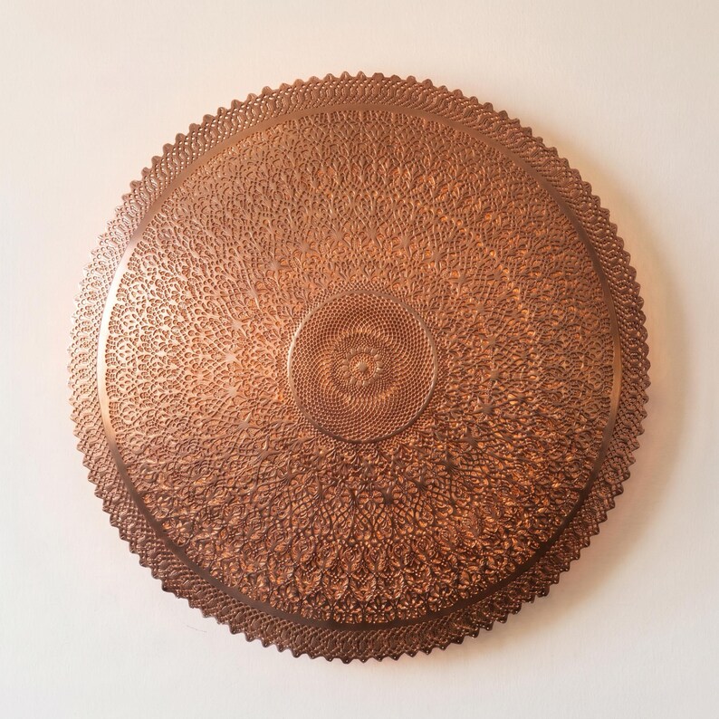 May include: A circular copper-colored wall plaque featuring an intricate, embossed pattern of repeating geometric and floral motifs. The design radiates from a central point toward a scalloped, serrated edge, creating a textured, metallic surface.