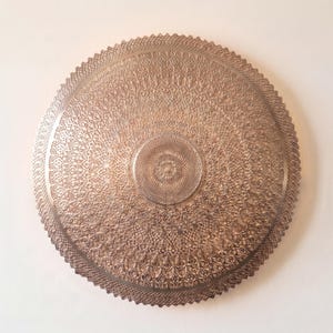 May include: A circular metallic wall plaque featuring a detailed, intricate lace-like pattern. The design consists of concentric rings of fine, repetitive geometric cutouts and textures, finished in a warm, muted bronze or copper tone.