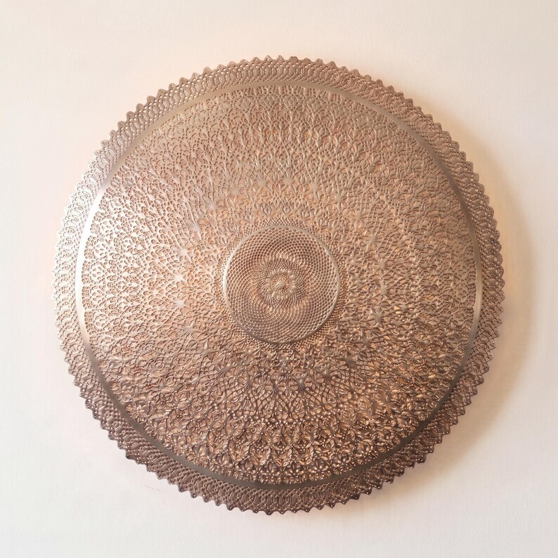 May include: A circular metallic wall plaque featuring a detailed, intricate lace-like pattern. The design consists of concentric rings of fine, repetitive geometric cutouts and textures, finished in a warm, muted bronze or copper tone.