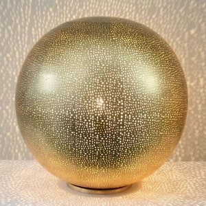 May include: A gold sphere lamp with a patterned surface. The lamp is illuminated, casting a pattern of light on the surface below.