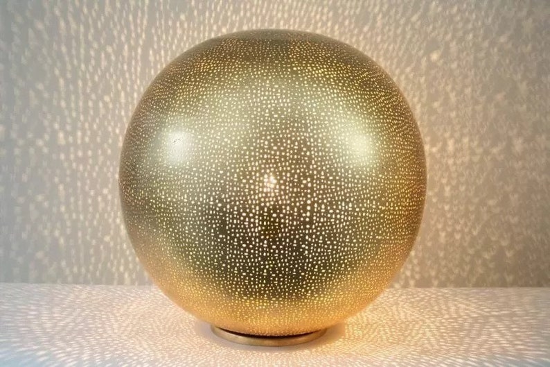 May include: A gold sphere lamp with a patterned surface. The lamp is illuminated, casting a pattern of light on the surface below.