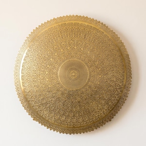 May include: A circular gold-colored metal wall plaque featuring an intricate, laser-cut filigree pattern. The design consists of a dense, repeating geometric motif radiating from a central point toward a scalloped, serrated outer edge.