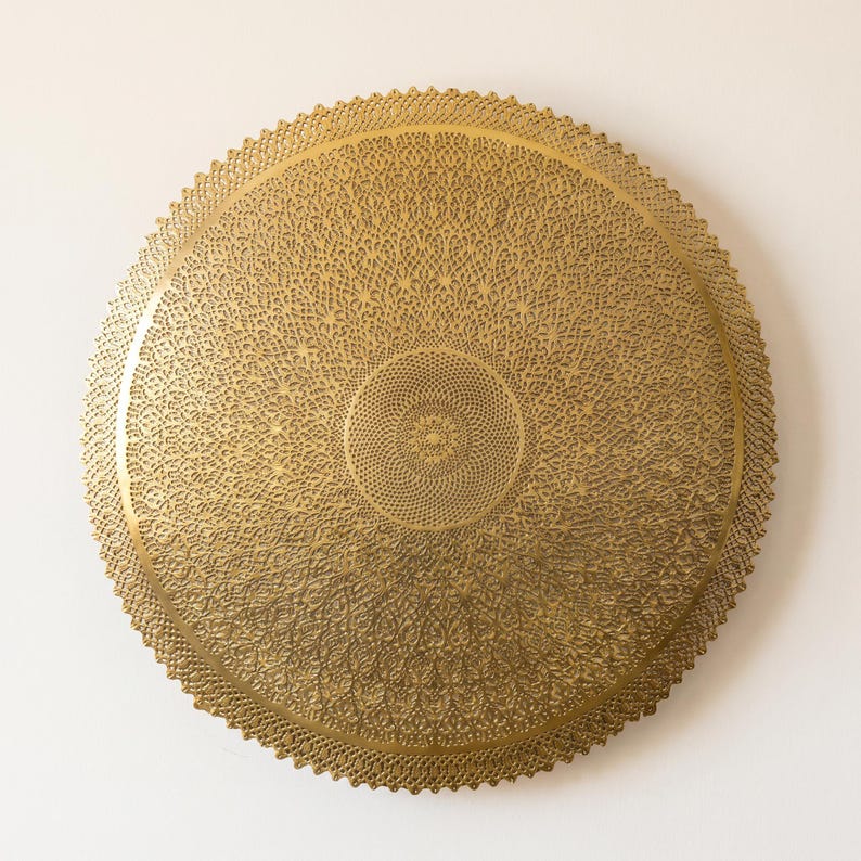 May include: A circular gold-colored metal wall plaque featuring an intricate, laser-cut filigree pattern. The design consists of a dense, repeating geometric motif radiating from a central point toward a scalloped, serrated outer edge.