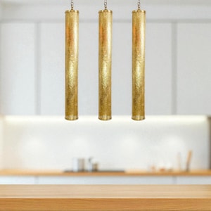 May include: Three gold cylindrical pendant lights hanging from the ceiling. The lights have a textured surface and are suspended above a wooden countertop. The kitchen has white cabinets and a stainless steel oven.
