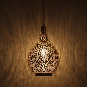 May include: A teardrop-shaped metal pendant light fitting with an intricate laser-cut pattern that emits a warm, glowing light. The lamp features a dark, metallic finish and is suspended from a thin chain, casting detailed shadows on the wall.