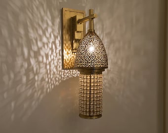 May include: A brass wall sconce with a decorative, patterned shade and a crystal-studded base. The sconce is mounted on a rectangular backplate and casts a patterned light on the wall. The overall design is ornate and elegant.