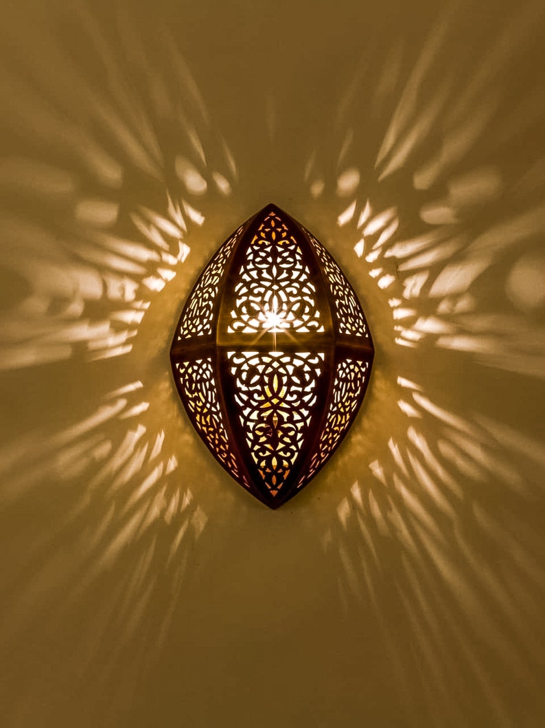 May include: A wall-mounted sconce with an elongated, pointed oval shape made of dark metal. The surface features intricate, laser-cut filigree patterns that allow warm, golden light to shine through, casting dramatic, radiating shadows on the wall.