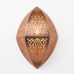 May include: A copper-colored wall sconce with an elongated, pointed oval shape. The metal surface features intricate, laser-cut filigree patterns that allow warm light to shine through, creating a glowing effect across the detailed cutouts.