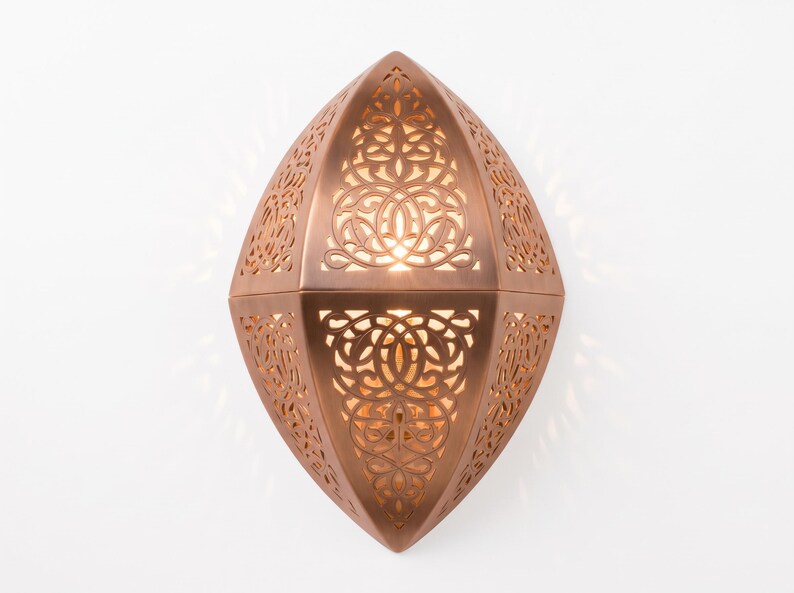 May include: A copper-colored wall sconce with an elongated, pointed oval shape. The metal surface features intricate, laser-cut filigree patterns that allow warm light to shine through, creating a glowing effect across the detailed cutouts.