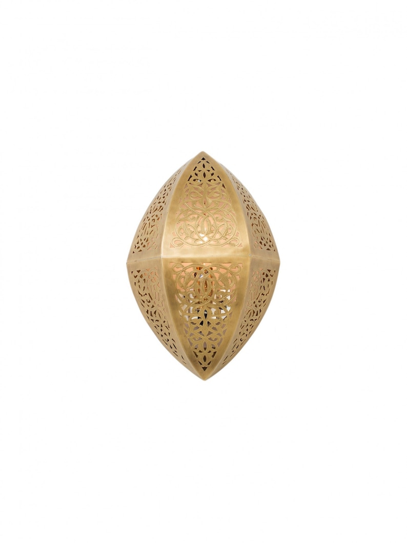 May include: A gold-colored metal wall sconce with an elongated, pointed oval shape. The surface features intricate, laser-cut filigree patterns throughout, allowing light to pass through the decorative cutouts in a symmetrical, ornate design.