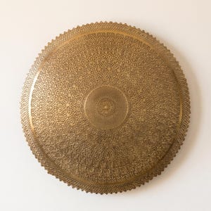 May include: A circular gold-toned metal tray featuring an intricate, laser-cut filigree pattern that radiates from a dense central point toward a scalloped, serrated edge. The surface displays a uniform metallic finish with detailed geometric cutouts.