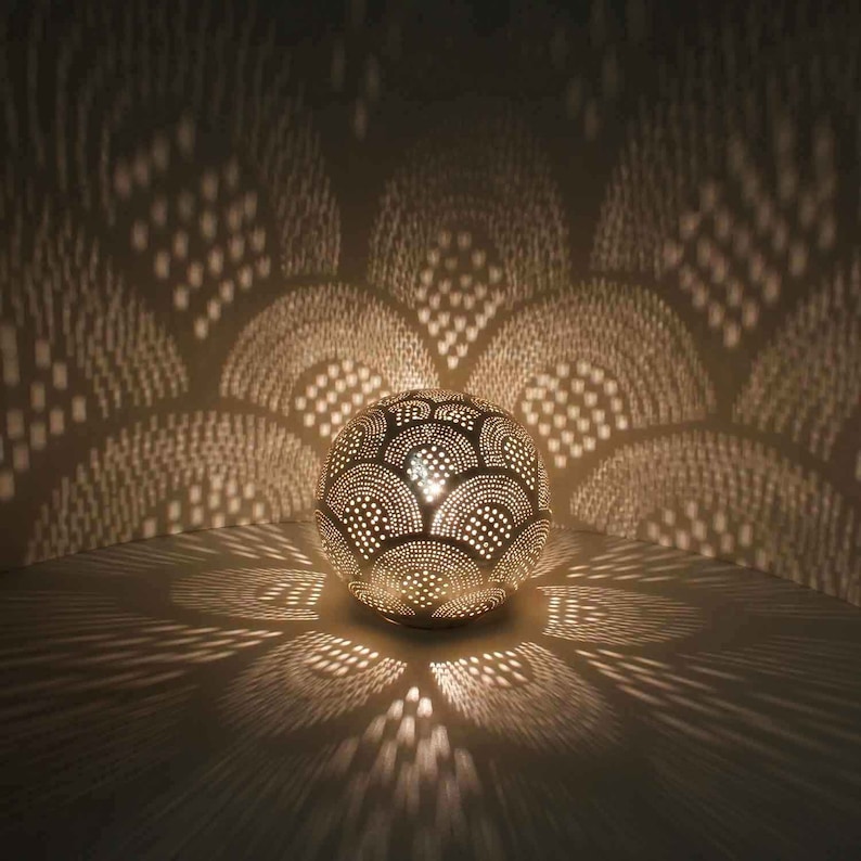 May include: A white metal lamp with a round, perforated shade that casts intricate patterns of light on the wall. The shade is decorated with a repeating pattern of overlapping circles.