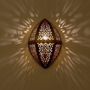 May include: A wall-mounted sconce with an almond-shaped metal frame featuring intricate, laser-cut filigree patterns. The fixture emits a warm, glowing light through the cutouts, casting dramatic, symmetrical shadows onto the surrounding wall surface.