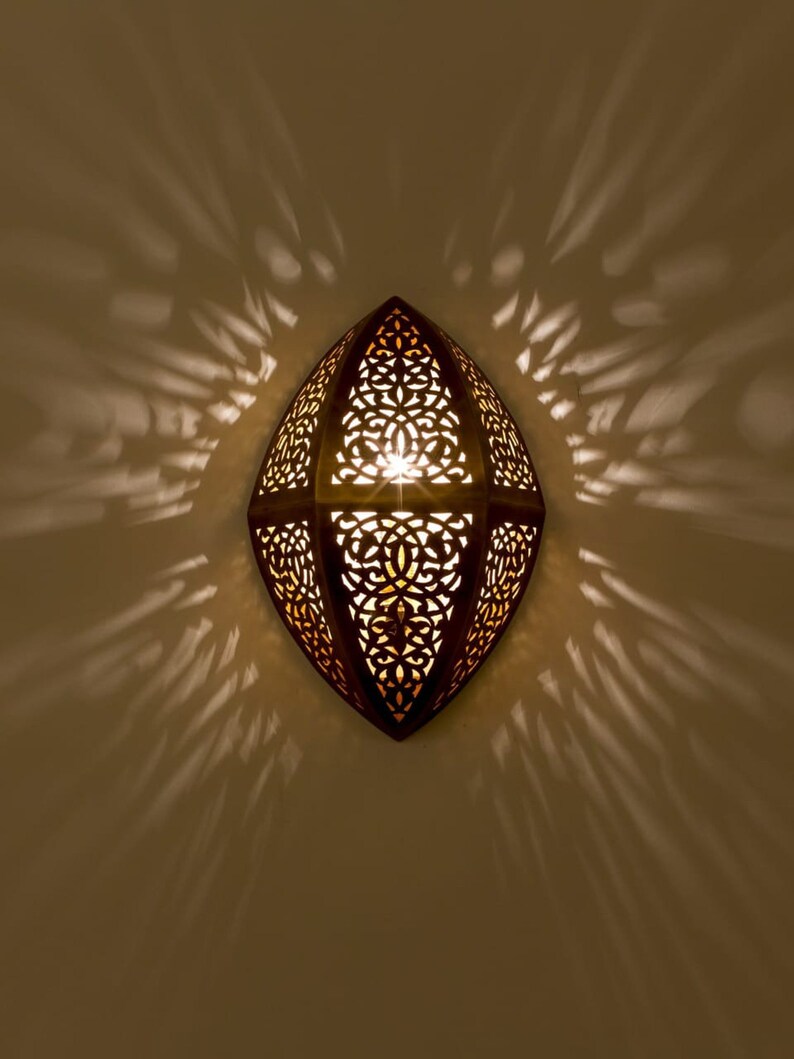 May include: A wall-mounted sconce with an almond-shaped metal frame featuring intricate, laser-cut filigree patterns. The fixture emits a warm, glowing light through the cutouts, casting dramatic, symmetrical shadows onto the surrounding wall surface.
