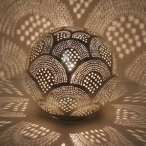 May include: A silver metal globe lamp with a decorative pattern of circles and arches. The lamp is lit and casting a warm glow.