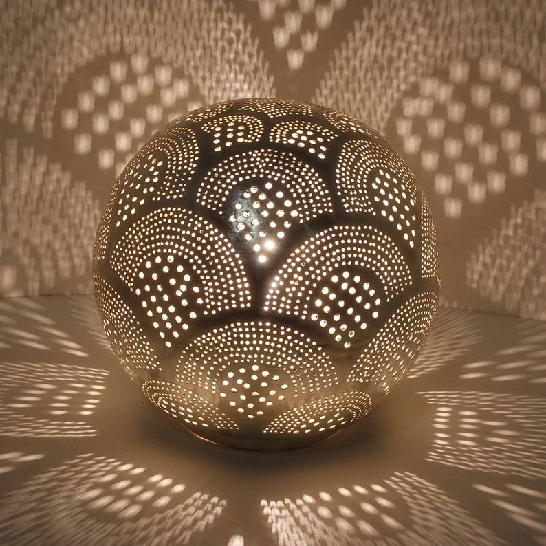 May include: A silver metal globe lamp with a decorative pattern of circles and arches. The lamp is lit and casting a warm glow.