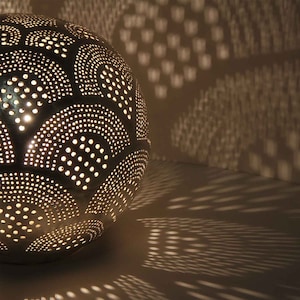 May include: A silver metal globe lamp with a decorative pattern of circles and arches. The lamp is lit and casting a shadow on the wall behind it.