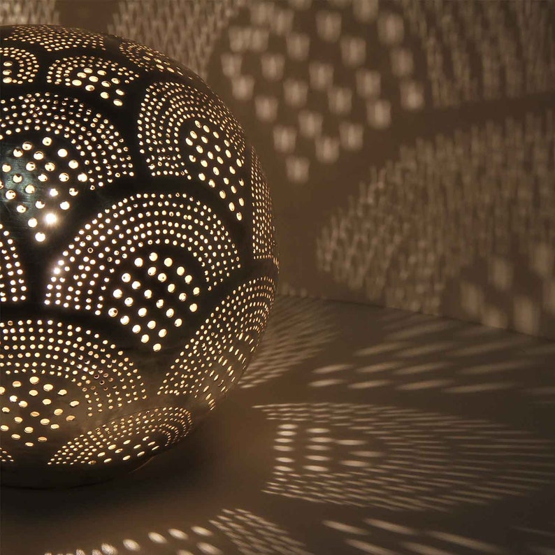 May include: A silver metal globe lamp with a decorative pattern of circles and arches. The lamp is lit and casting a shadow on the wall behind it.