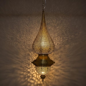 May include: A teardrop-shaped brass pendant light fixture featuring intricate laser-cut patterns that cast detailed shadows. The metal lamp has a pointed top, a wider central body, and a smaller rounded base, all suspended by a thin metal chain.