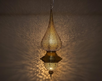 May include: A teardrop-shaped brass pendant light fixture featuring intricate laser-cut patterns that cast detailed shadows. The metal lamp has a pointed top, a wider central body, and a smaller rounded base, all suspended by a thin metal chain.