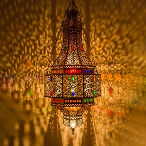 May include: A gold metal Moroccan lantern with intricate cutouts and colorful glass accents. The lantern is hanging from a chain and casts intricate shadows on the wall.