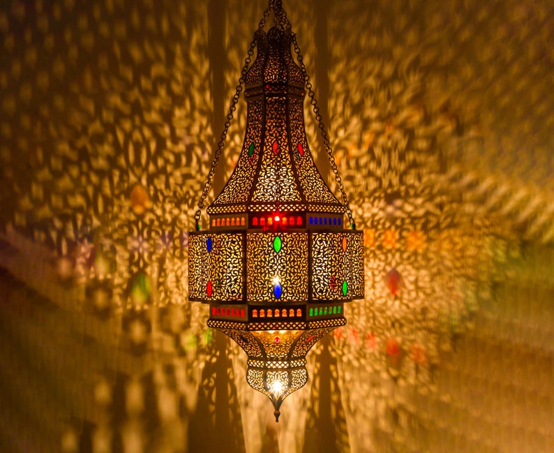May include: A gold metal Moroccan lantern with intricate cutouts and colorful glass accents. The lantern is hanging from a chain and casts intricate shadows on the wall.