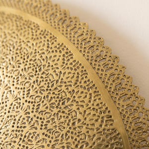May include: A circular gold-colored metal tray featuring an intricate, laser-cut filigree pattern across its surface. The design consists of dense, repeating floral and geometric motifs with a scalloped, lace-like edge along the outer rim.