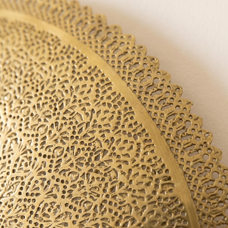 May include: A circular gold-colored metal tray featuring an intricate, laser-cut filigree pattern across its surface. The design consists of dense, repeating floral and geometric motifs with a scalloped, lace-like edge along the outer rim.