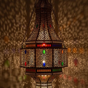 May include: A large, ornate, metal lantern with intricate cutouts and colorful glass accents. The lantern is hanging from a chain and is illuminated from within, casting intricate shadows on the wall.