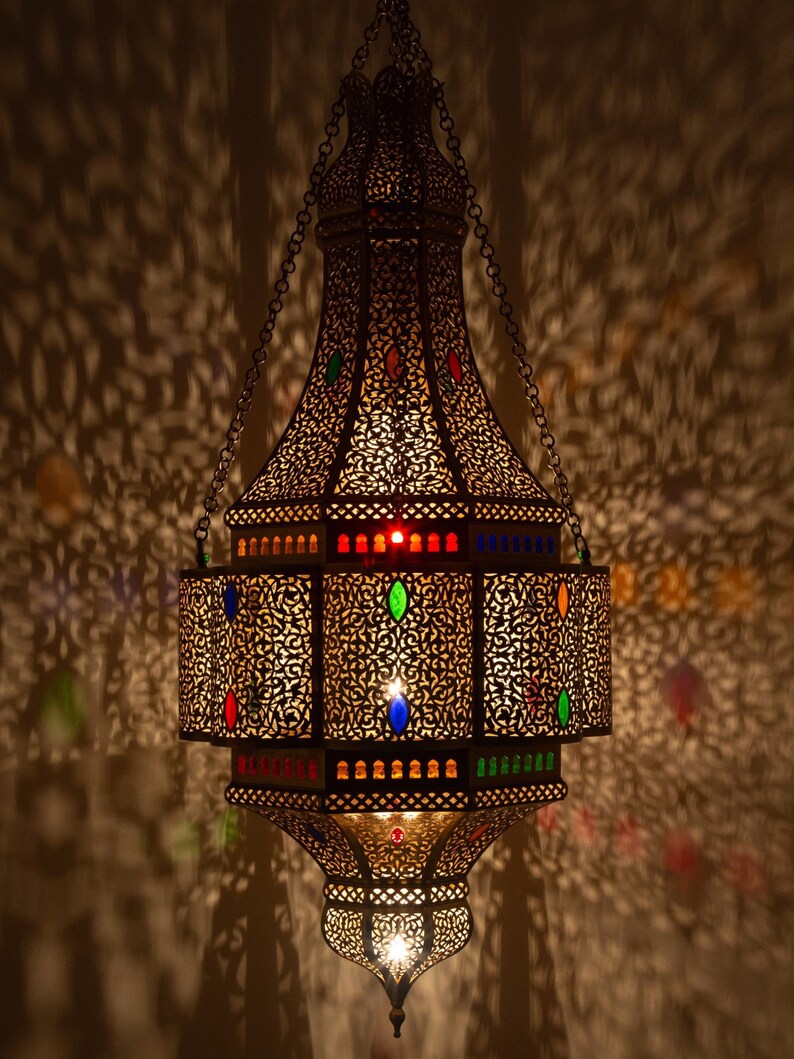 May include: A large, ornate, metal lantern with intricate cutouts and colorful glass accents. The lantern is hanging from a chain and is illuminated from within, casting intricate shadows on the wall.