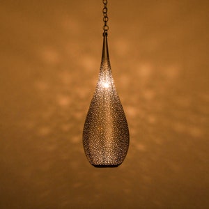 Pode incluir: Uma luminária pendente em forma de lágrima prateada com um padrão delicado e intrincado. A luz está pendurada em uma corrente contra um fundo marrom quente.