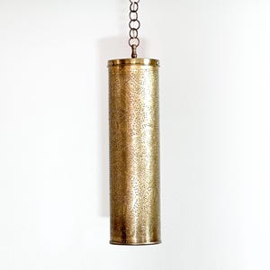 May include: A cylindrical pendant light fixture with a brass finish. The light has a detailed, perforated pattern across its surface. It hangs from a chain, and the fixture is suspended against a white background.