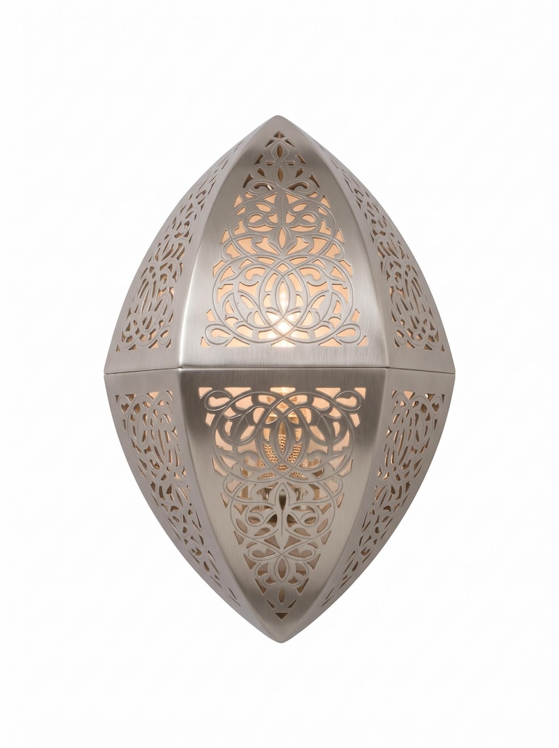 May include: A brushed nickel wall sconce with an elongated, pointed oval shape. The metal fixture features intricate, laser-cut filigree patterns that allow warm light to glow from within, creating a decorative effect on the surface.