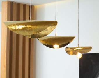 May include: Three gold-toned pendant lights with hammered metal shades in a crescent shape. The lights are hanging from the ceiling and are illuminated.