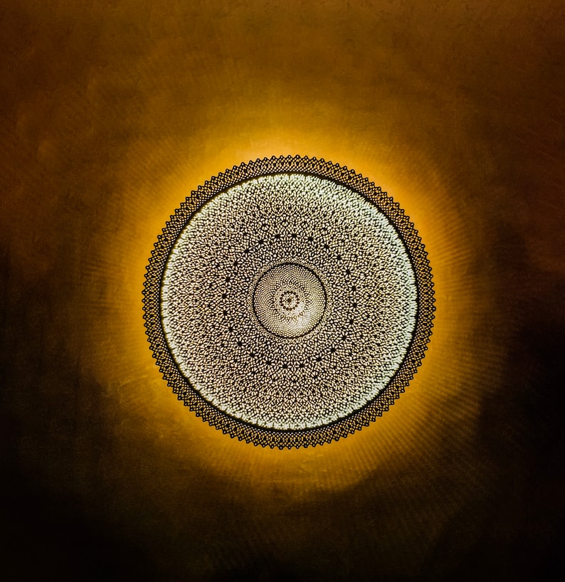 May include: A circular wall lamp featuring an intricate, dark metal lattice design with a fine, repeating geometric pattern. The light source behind the metal structure emits a warm, golden glow that radiates outward, creating a soft halo effect.