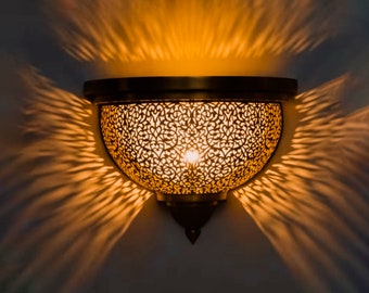 May include: A brass wall sconce with a decorative, intricate, cut-out design. The sconce is lit and casts a warm, golden glow on the wall.