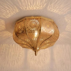 May include: A gold-toned metal flush mount ceiling light fixture with an intricate, perforated pattern of circular and geometric cutouts. The light emits a warm glow, casting detailed, radiating shadow patterns onto the surrounding ceiling surface.