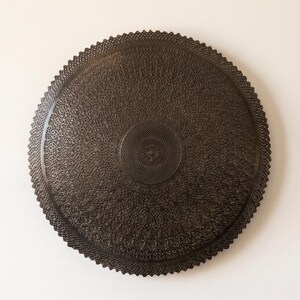 May include: A circular dark brown wall plaque featuring an intricate, laser-cut lace-like pattern. The design consists of dense, repeating geometric motifs radiating from a central point toward a scalloped, serrated outer edge.