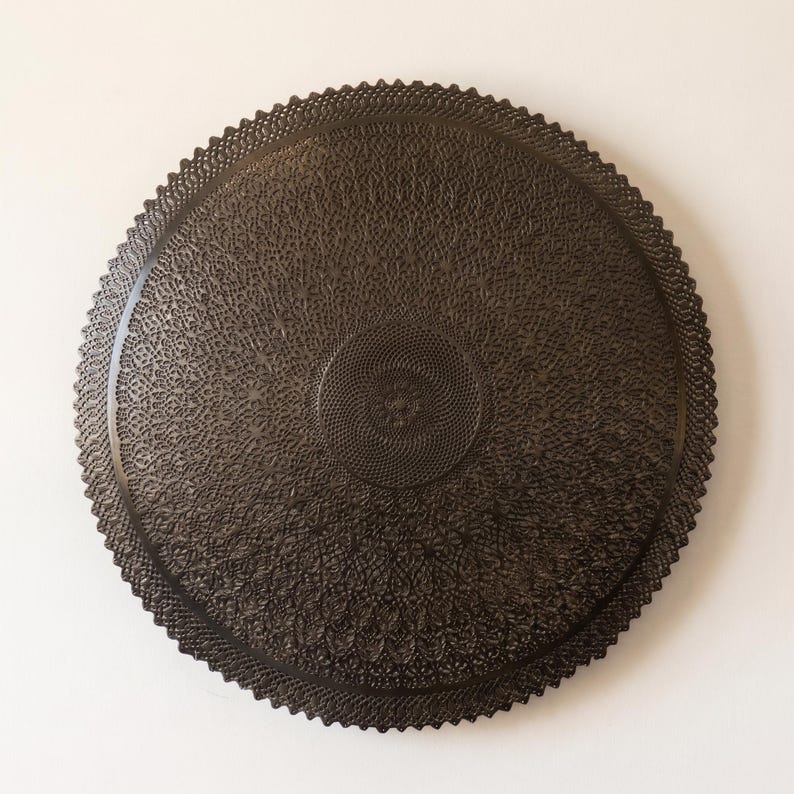 May include: A circular dark brown wall plaque featuring an intricate, laser-cut lace-like pattern. The design consists of dense, repeating geometric motifs radiating from a central point toward a scalloped, serrated outer edge.
