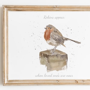 Puede incluir: Una impresión en acuarela enmarcada con un petirrojo posado en un poste de madera. El ave tiene el pecho rojo y está rodeada de salpicaduras de pintura. El texto "Robins appear when loved ones are near" está escrito arriba y abajo.