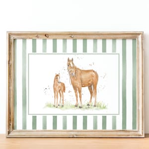 May include: A framed watercolor print featuring a brown mare and her foal standing in green grass. The artwork is centered on a white background with a border of vertical sage green and white stripes, all encased in a light wood frame.